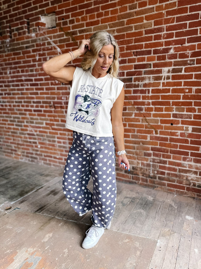 A woman posing in front of a red brick wall wearing a white sleeveless graphic crop top and purple wide-leg pants with white polka dots, and white sneakers.