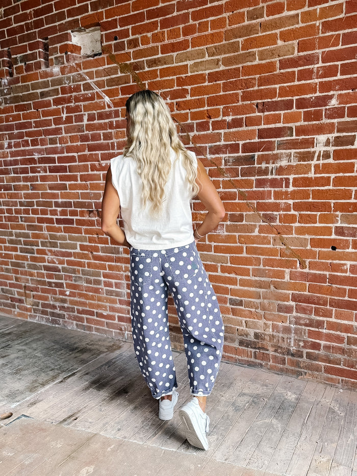 A woman with long blonde hair is shown from behind, wearing loose-fitting purple pants with white polka dots, a white sleeveless top, and white sneakers, standing in front of a red brick wall