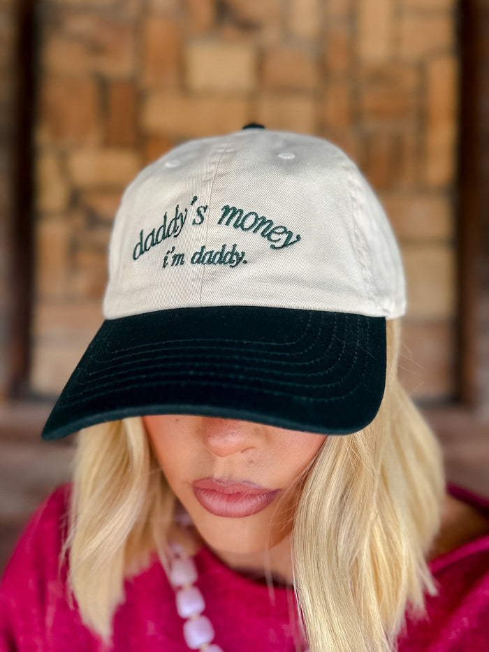 A woman wearing a two-tone baseball cap with a cream crown and a black brim. The cap features the embroidered text 'daddy's money i'm daddy.