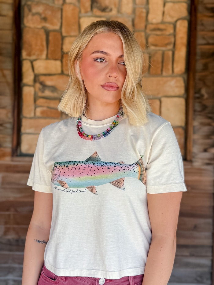 A blonde woman wearing a white graphic t-shirt with a colorful rainbow trout print and the text 'I'd rather eat fish than meat', posed in front of a rustic stone wall.