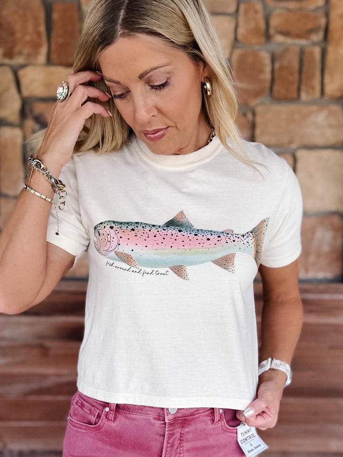 A woman wearing a white graphic t-shirt with a pink and blue trout design, paired with dusty rose jeans, standing in front of a rustic brick wall.