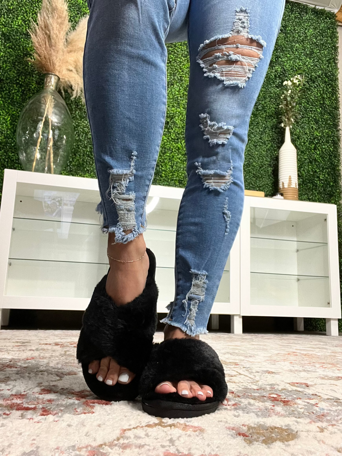 Black fur slippers with a hard sole, worn by a person standing on a rug.