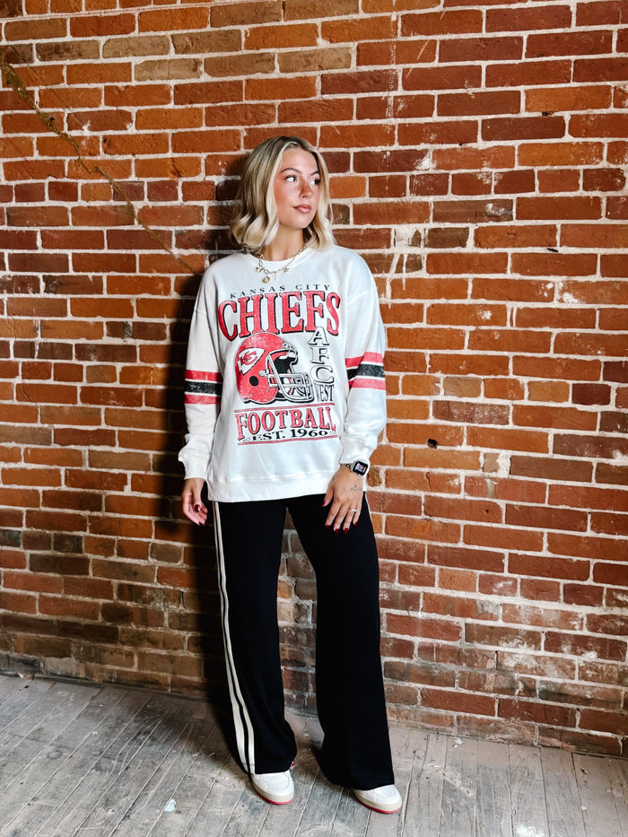 A woman stands against a red brick wall wearing a white long-sleeve graphic sweatshirt with red and navy lettering that reads 'Chiefs Football 2025' and black wide-leg track pants with white side stripes.