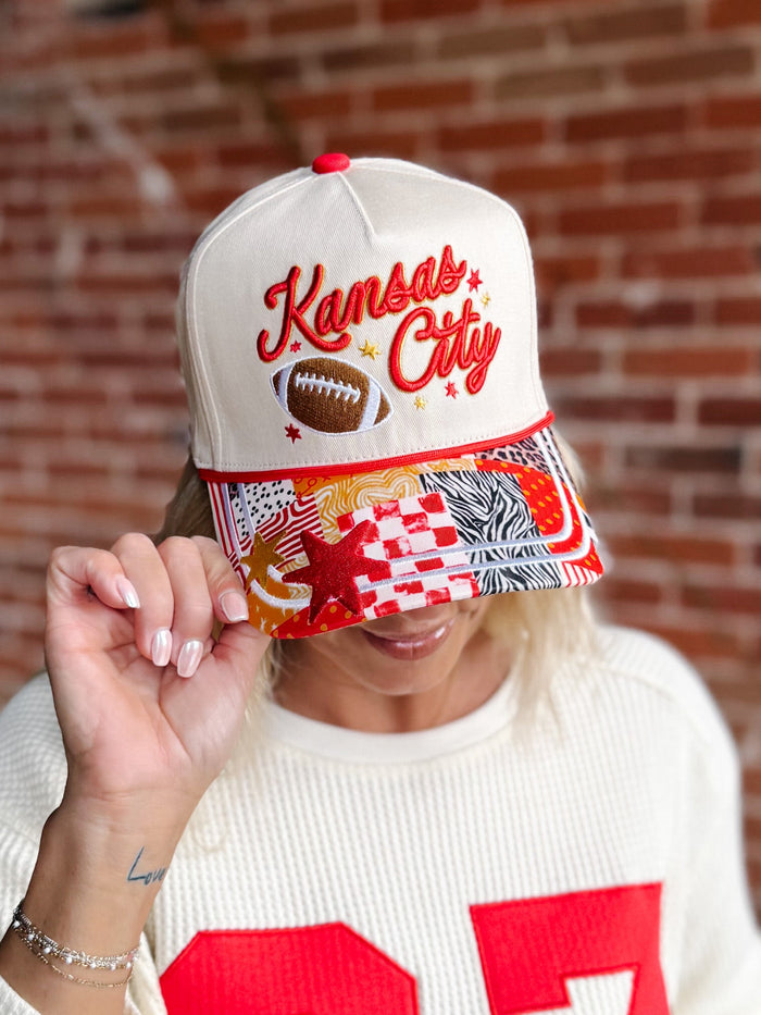 A person wearing a cream snapback cap with 'Kansas City' embroidered in red on the front, alongside a football graphic. The brim features a busy multicolor patchwork pattern with red, white, black, and brown elements.