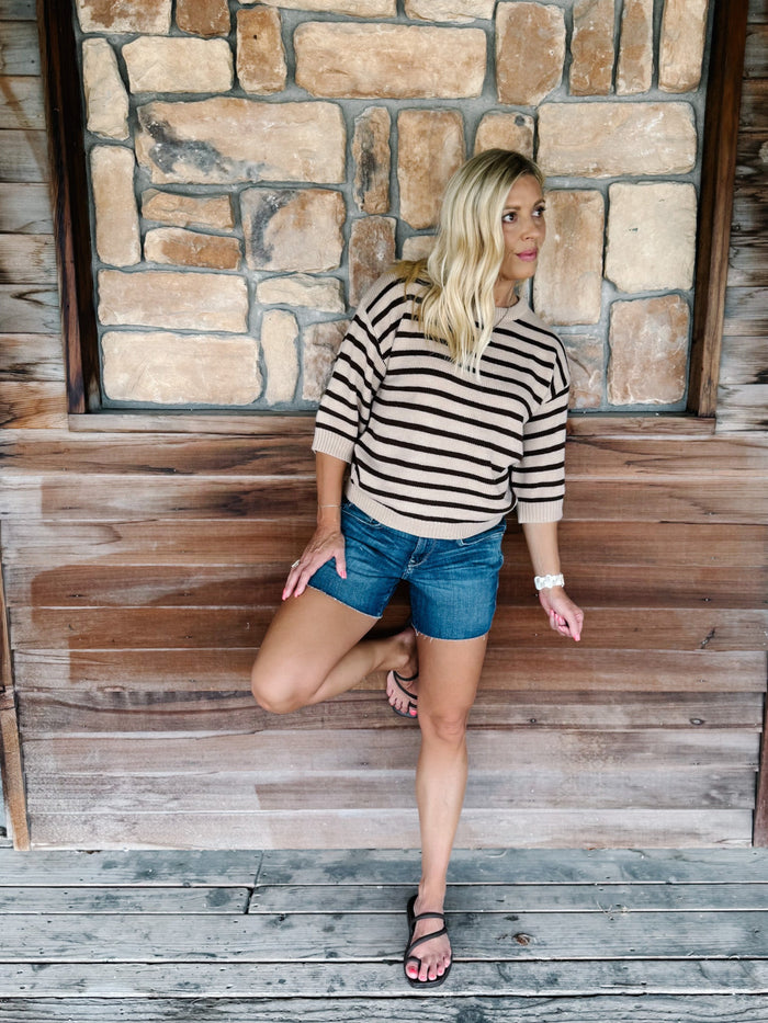 A woman posing on a rustic wooden porch with a stone wall background, wearing a beige and black striped long-sleeve knit top, denim shorts, and black sandals.