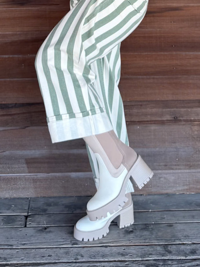 White platform Chelsea boots with chunky lug soles and elastic side panels, worn with green and white striped trousers against a rustic wooden plank background.