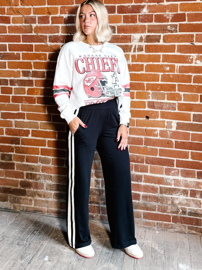 A woman stands against a red brick wall wearing a white graphic sweatshirt with red and pink 'CHIEFS' lettering and a football helmet print, paired with black wide-leg track pants featuring white side stripes, and white sneakers.