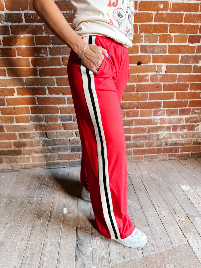 A person wearing bright red wide-leg track pants with a black and white side stripe, standing against a brick wall and wood floor.