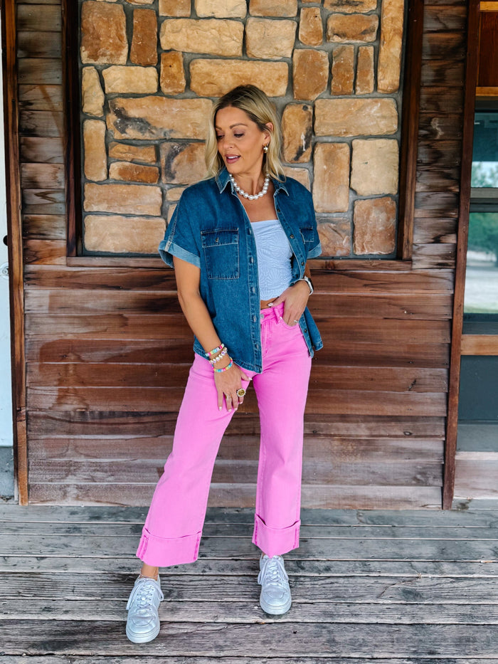 A woman stands on a wooden porch wearing bright pink wide-leg jeans, a light blue tank top, a dark blue denim shirt, and white sneakers, with a stone wall and wood door behind her.