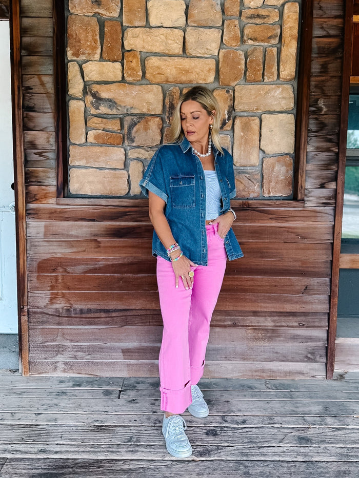 A woman stands on a rustic wooden porch in front of a stone wall, wearing a blue denim shirt, bright pink pants, and white sneakers.