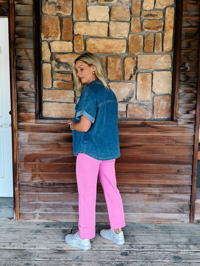 A woman stands on a wooden porch in front of a stone wall, wearing a blue denim shirt with rolled sleeves, bright pink pants, and white sneakers.