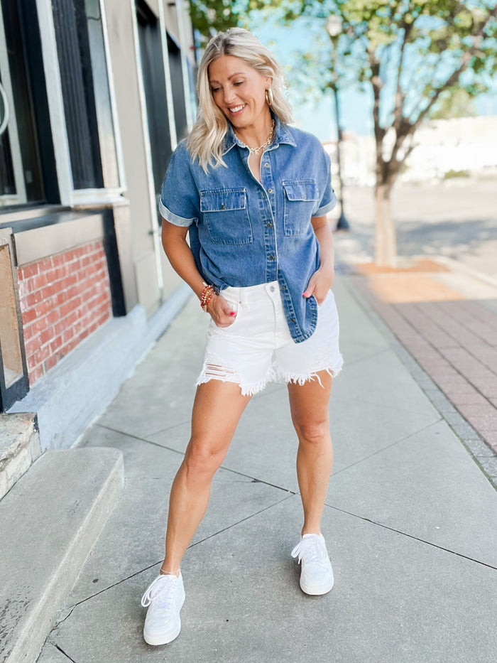 A woman stands on a sunny sidewalk wearing a short-sleeve button-up denim shirt, white frayed shorts, and white sneakers.