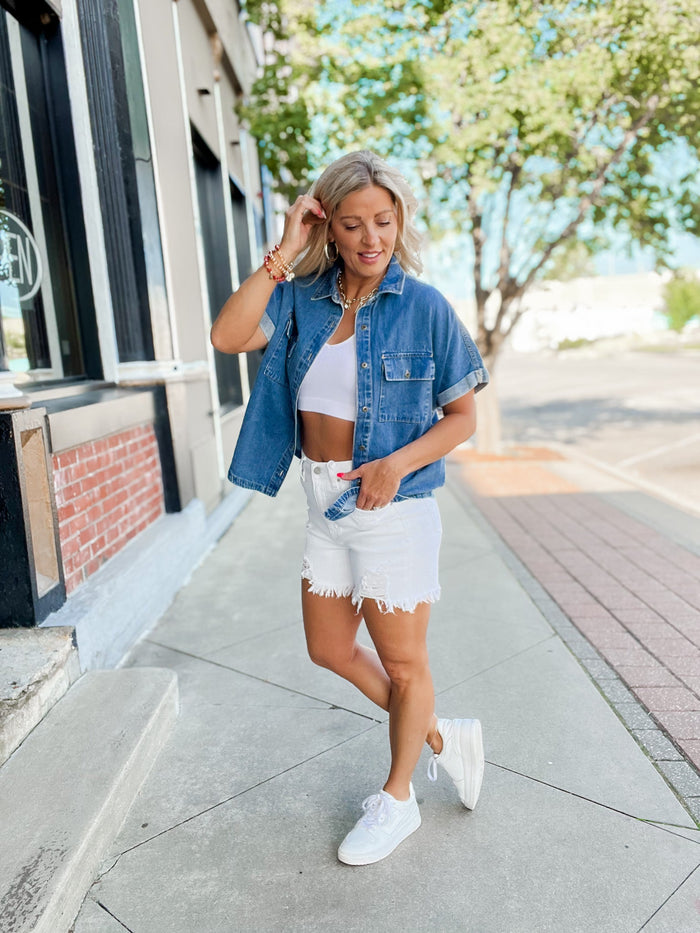 Woman in denim shirt and white shorts walking on a sidewalk.