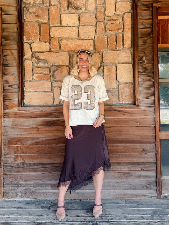 A woman stands on a rustic wooden porch in front of a stone wall, wearing a cream short-sleeve athletic-style top with a large beige '23' graphic and a dark brown midi skirt with a lace-trim hem.