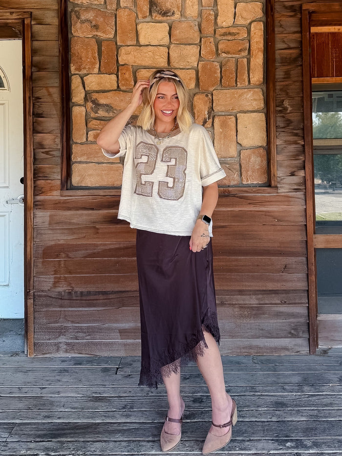 A woman stands on a rustic wooden porch in front of a stone wall, wearing a cream graphic t-shirt with a patchwork number 23 and a dark brown midi skirt.