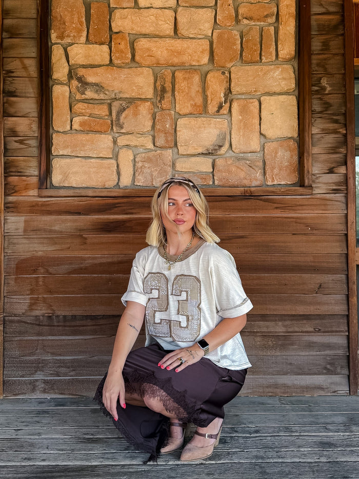 A woman crouching on a rustic wooden porch in front of a stone wall, wearing a white t-shirt with a glittery silver number 23, dark brown pants, and beige shoes.